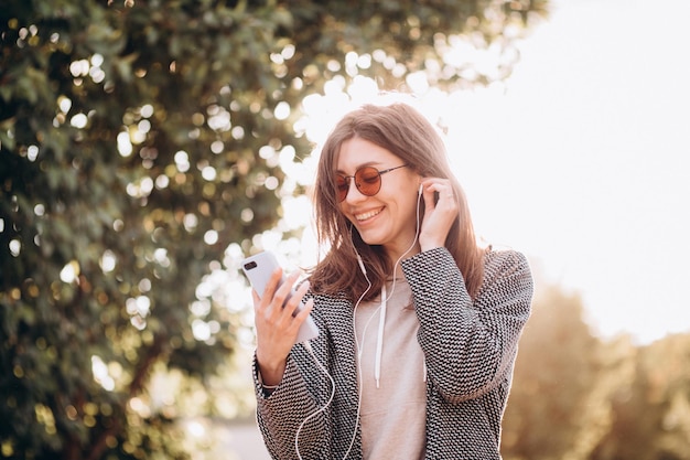 Belle jeune femme avec un sourire écoute et apprécie la musique du téléphone dans les écouteurs dans le parc