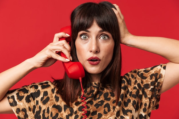 belle jeune femme surprise vêtue d'une chemise imprimée d'animaux posant isolée sur un mur rouge parlant par téléphone.