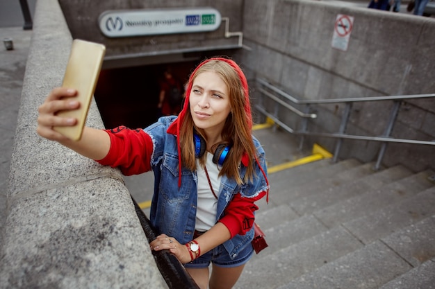 Belle jeune femme en sweat à capuche rouge prend selfie à l'aide d'un téléphone portable près de l'entrée du métro.