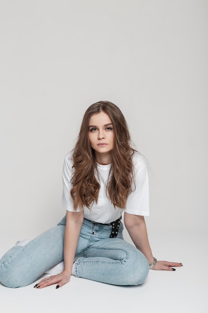 Belle jeune femme avec un t-shirt blanc et un jean vintage bleu est assise en studio sur fond blanc