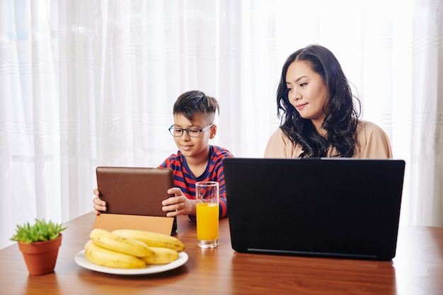 Belle jeune femme à la table de la cuisine travaillant sur ordinateur portable, son fils assis à proximité et regardant une vidéo éducative sur tablette numérique