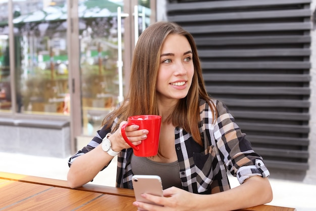 Belle jeune femme avec une tasse de boisson au cacao sucré à l'aide d'un téléphone portable au café