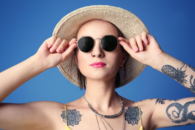 Belle jeune femme avec tatouage portant chapeau et lunettes de soleil sur fond bleu