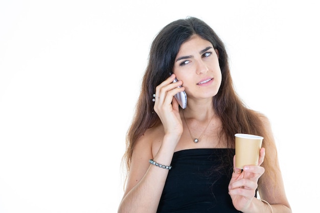 Belle jeune femme sur téléphone mobile tout en buvant une boisson chaude tasse de thé en carton de café sur fond blanc isolé