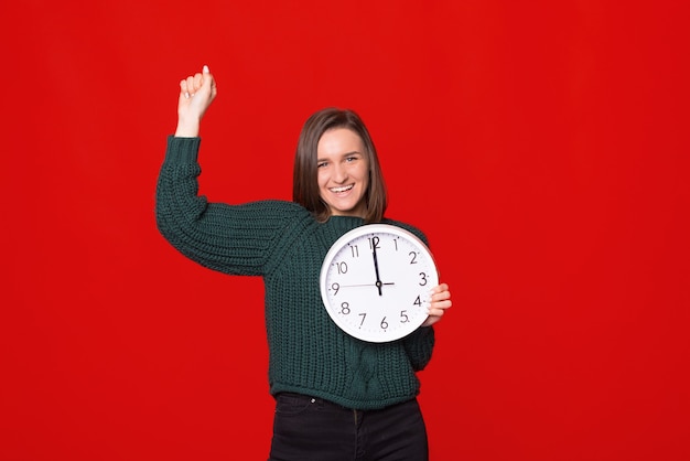 Belle jeune femme tenant une grande horloge célébrant la victoire avec un sourire heureux et l'expression du gagnant avec les mains levées