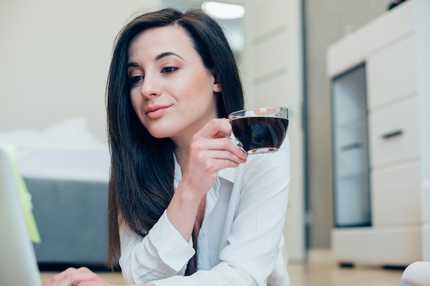 Belle jeune femme tenant une tasse de café en verre et regardant l'écran de l'ordinateur portable à la maison