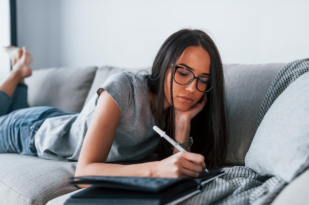 Belle jeune femme en tenue décontractée allongée seule à la maison avec un bloc-notes et un stylo.