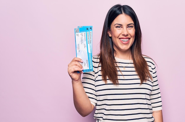 Belle jeune femme touristique tenant une carte d'embarquement de la compagnie aérienne sur fond rose isolé, l'air positif et heureux debout et souriant avec un sourire confiant montrant les dents