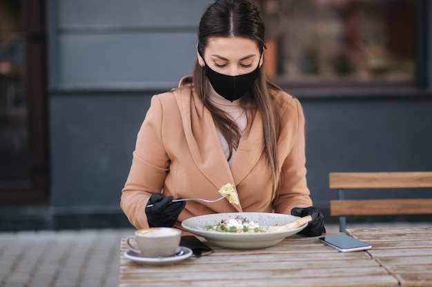 Belle jeune femme en train de déjeuner au café en plein air sur la terrasse. Femme en masque de protection noir.