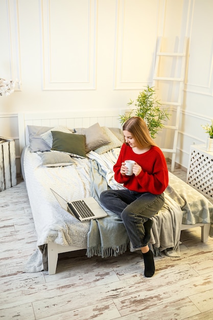 Belle jeune femme travaillant sur un ordinateur portable assise sur le lit à la maison, elle boit du café et sourit. Travail à domicile pendant le confinement. Fille dans un pull et un jean à la maison sur le lit