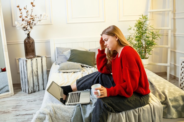 Belle jeune femme travaillant sur un ordinateur portable assise sur le lit à la maison, elle boit du café et sourit. Travail à domicile pendant le confinement. Fille dans un pull et un jean à la maison sur le lit