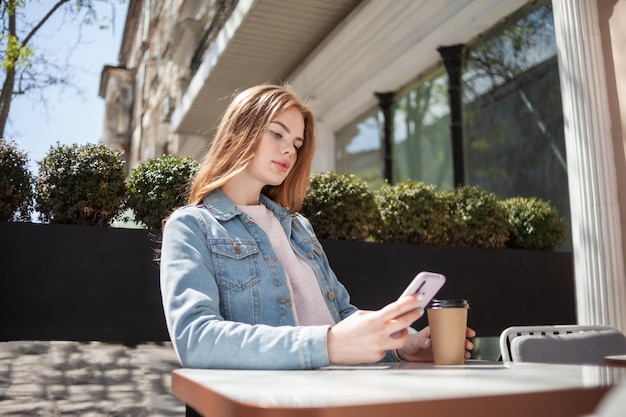 Belle jeune femme utilisant un smartphone et buvant du café assis à une table dans un café en plein air