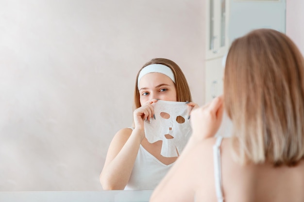 Belle jeune femme utilise un masque facial en feuille sur la peau du visage pour les soins de la peau du spa de beauté à la salle de bain à domicile