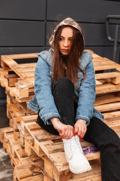 Belle jeune femme en veste en jean à la mode en jeans en bottes de cuir posant sur les planches dans la rue. Mannequin fille élégante reposant sur des palettes en bois dans la ville. Style décontracté pour les jeunes.