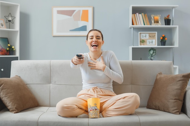 Belle jeune femme en vêtements décontractés assis sur un canapé à l'intérieur de la maison avec un seau de pop-corn tenant à distance regarder un film heureux et excité en riant passer du temps à la maison