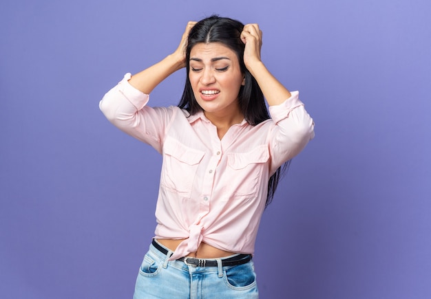 Belle Jeune Femme En Vêtements Décontractés, Ennuyée Et Irritée, Touchant Sa Tête Avec Les Mains Debout Sur Le Mur Bleu