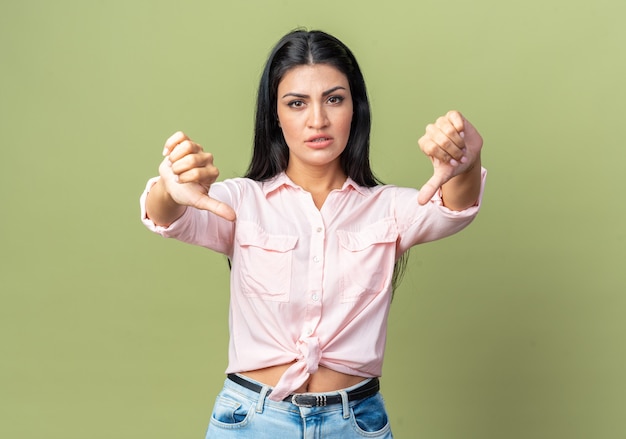 Belle Jeune Femme En Vêtements Décontractés étant Mécontente Montrant Les Pouces Vers Le Bas Debout Sur Le Mur Vert