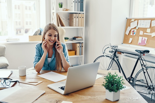 Belle jeune femme en vêtements décontractés intelligents souriant et parlant au téléphone alors qu'elle était assise au bureau