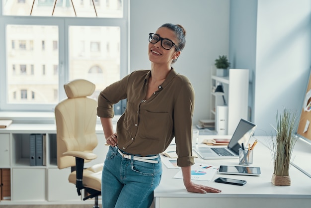 Belle jeune femme en vêtements décontractés intelligents souriant tout en se tenant au bureau