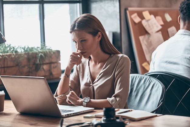 Belle jeune femme en vêtements décontractés intelligents utilisant un ordinateur portable tout en travaillant au bureau