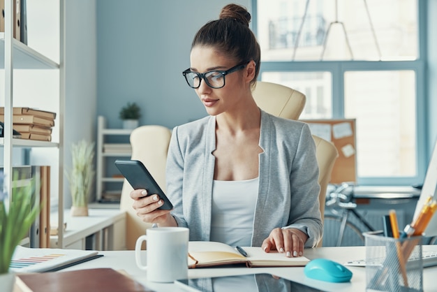 Belle jeune femme en vêtements décontractés intelligents utilisant un téléphone et souriant tout en travaillant au bureau