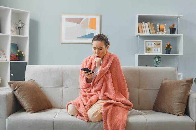 Belle jeune femme en vêtements de maison assise sur un canapé sous une couverture à l'intérieur de la maison se sentant mal à l'aide d'un smartphone toussant attrapant un rhume