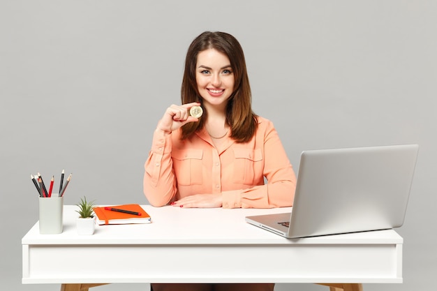 Belle jeune femme en vêtements pastel tenant bitcoin, future monnaie assis au bureau avec ordinateur portable pc isolé sur fond gris. Concept de style de vie de carrière d'affaires de réalisation. Maquette de l'espace de copie.