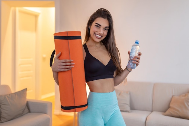 Belle jeune femme en vêtements de sport tient un tapis de yoga et une bouteille d'eau regardant la caméra et souriant