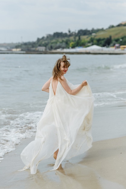 Une belle jeune femme vêtue d'une longue robe blanche se promène le long de la plage et de la jetée contre la mer.