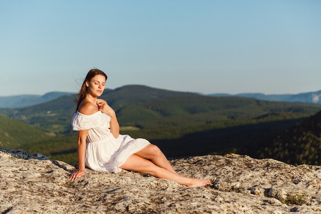 Belle jeune femme vêtue d'une robe blanche bénéficie de superbes paysages dans les montagnes au coucher du soleil