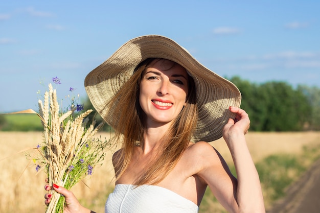 Belle jeune femme vêtue d'une robe blanche et d'un chapeau tient un bouquet de fleurs sauvages sur la route entre le champ laiteux.