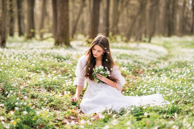 Belle jeune femme vêtue d'une robe blanche recueille des primevères Une fille dans la forêt de printemps Un bouquet d'anémones blanches dans ses mains