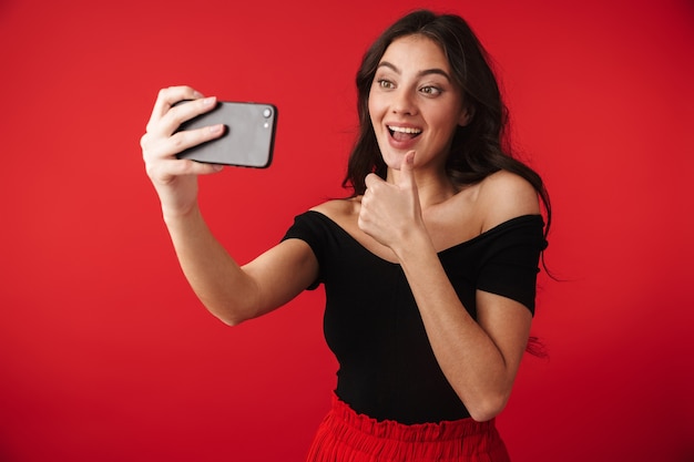 Belle jeune femme vêtue d'une robe debout isolé sur rouge, prenant un selfie