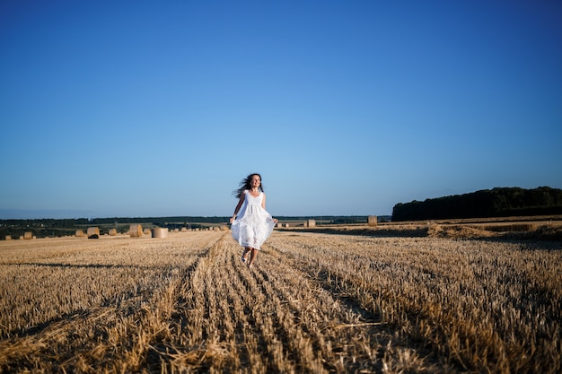 Une belle jeune femme vêtue d'une robe d'été blanche se dresse sur un champ de blé fauché avec d'énormes gerbes de foin, profitant de la nature. Nature au village. Mise au point sélective