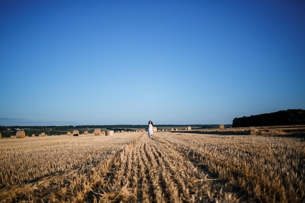 Une belle jeune femme vêtue d'une robe d'été blanche se dresse sur un champ de blé fauché avec d'énormes gerbes de foin, profitant de la nature. Nature au village. Mise au point sélective