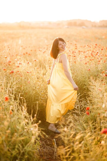 Belle jeune femme vêtue d'une robe jaune marchant dans un champ de coquelicots un jour d'été. Fille profitant des fleurs à la campagne. Mise au point sélective