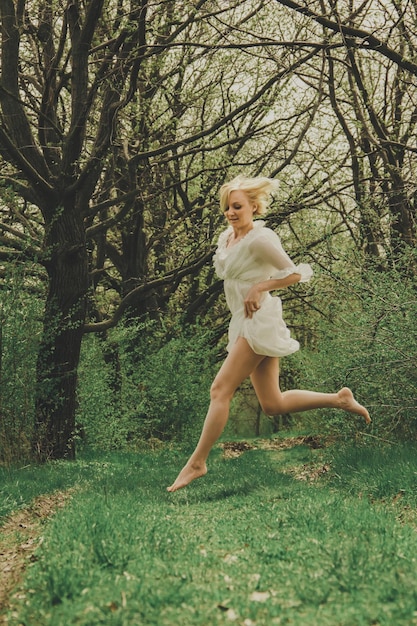 Une belle jeune femme vêtue d'une robe en mousseline blanche avec une courte coupe de cheveux blonde court le long du chemin et saute dans le parc.