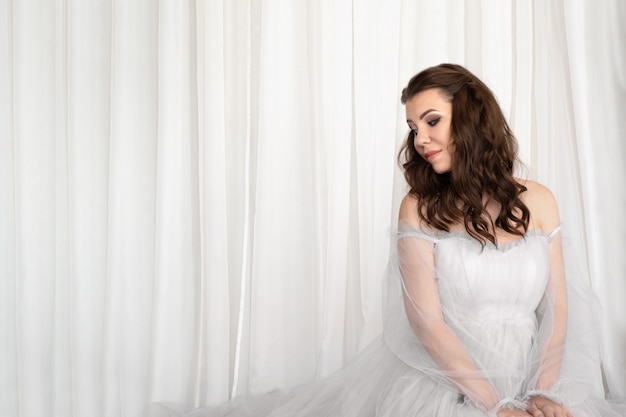 Belle jeune femme vêtue d'une robe romantique blanche assise sur un sol sur fond de tissu blanc.