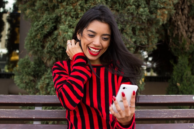 Belle jeune femme vêtue de rouge, assise sur un banc dans le parc et regardant son téléphone portable, souriant et célébrant la bonne nouvelle. Concept de vie moderne adolescent.