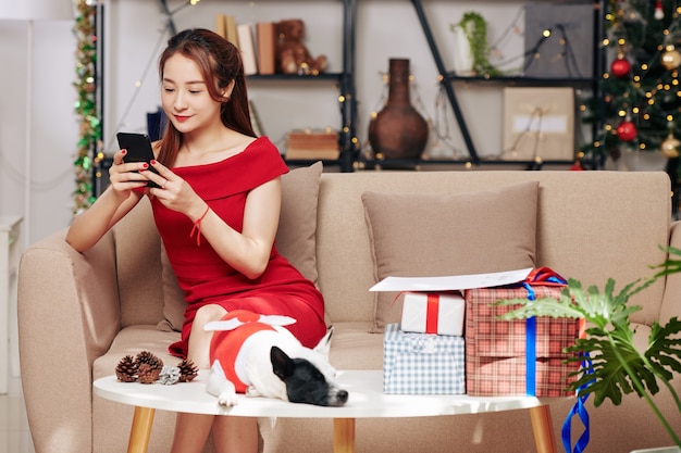 Belle jeune femme vietnamienne assise sur un canapé avec un petit chien en costume de Noël et une pile de cadeaux sur la table devant elle et vérifiant son smartphone