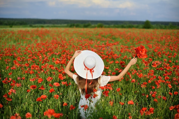 Belle jeune femme vivant le style tient dans les mains chapeau blanc et se tient à bras ouverts sur le champ de coquelicots rouges.