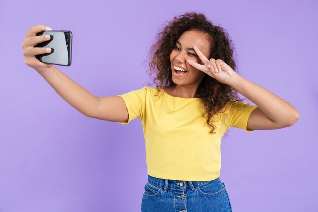 Belle jeune fille africaine heureuse portant des vêtements décontractés, isolée sur un mur violet, prenant un selfie, montrant un geste de paix