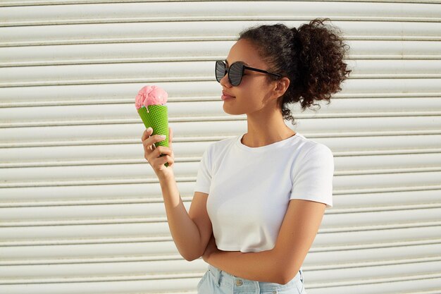 Une belle jeune fille afro-américaine dans un t-shirt blanc et un jean clair mange de la glace contre un mur clair par une journée ensoleillée