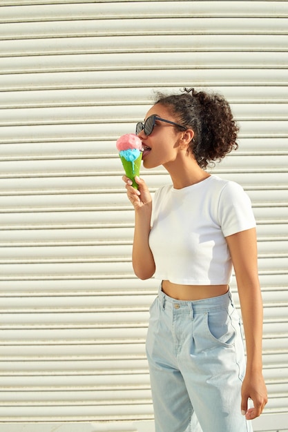 Une belle jeune fille afro-américaine dans un t-shirt blanc et un jean clair mange de la glace contre un mur clair par une journée ensoleillée