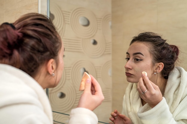 Belle jeune fille applique le fond de teint sur son visage au miroir dans la salle de bain Concept de maquillage au miroir