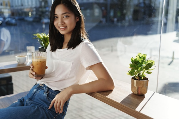 Belle jeune fille asiatique au café urbain profiter d'une boisson au café tenir un verre avec un comptoir maigre près de la fenêtre profiter d'une journée ensoleillée après l'université caméra souriante passer des loisirs endroit préféré