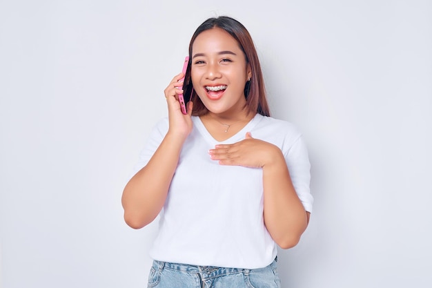 Belle jeune fille asiatique souriante portant un t-shirt blanc décontracté parle sur un téléphone portable en regardant la caméra avec une expression confiante et heureuse isolée sur fond blanc Concept de style de vie des gens