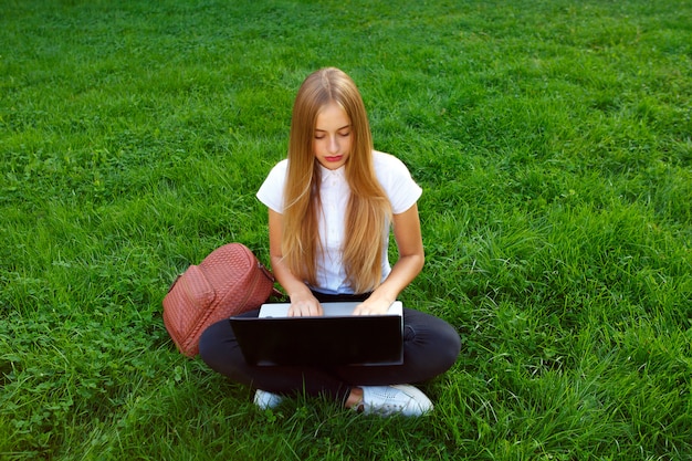 Belle jeune fille assise dans un parc sur l&#39;herbe verte avec un ordinateur portable