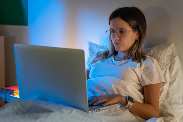 Une belle jeune fille assise sur le lit la nuit travaillant avec son ordinateur portable au milieu de la nuit