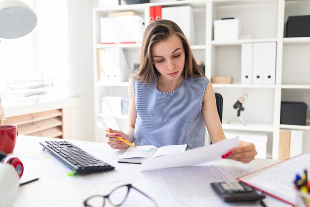 Belle jeune fille au bureau est assise à une table et travaille avec un crayon, un bloc-notes et des documents.